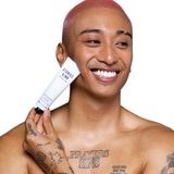 A person with pink hair smiles, holding a tube labeled "STORIES INK TATTOO CARE AFTERCARE CREAM" against their face. They have tattoos on their chest and hand, against a white background.